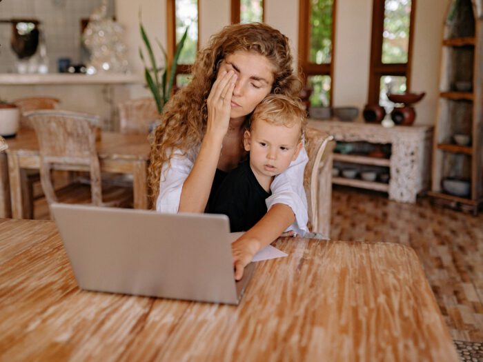 A woman with a baby on her knee looking at a laptop on a table and looking stressed